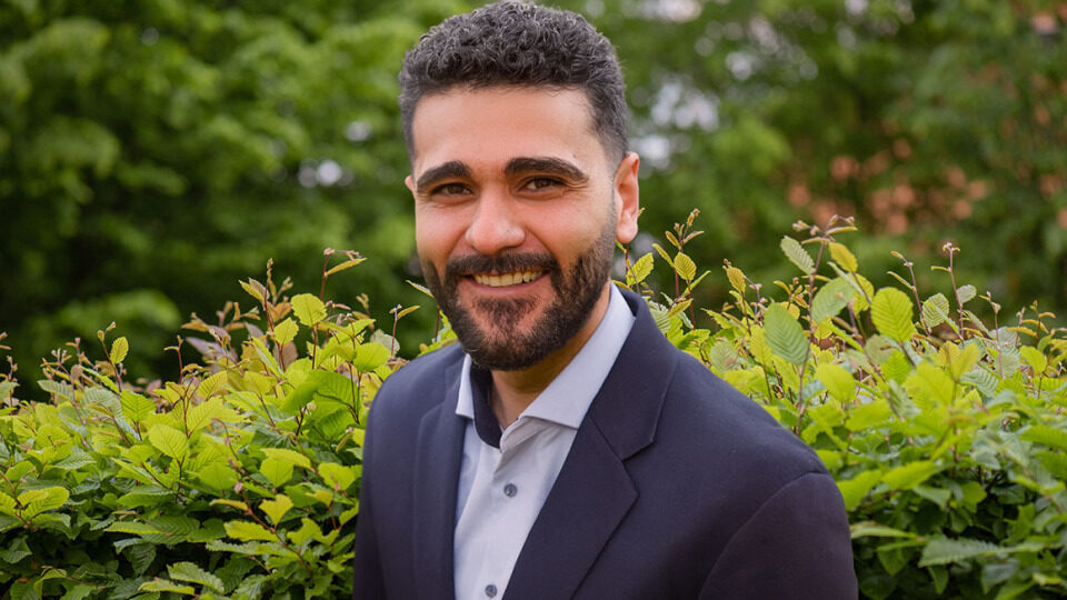 Projektleiter Uygar Celik steht vor einer grünen Hecke.